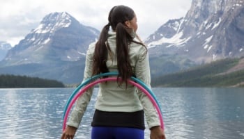 woman with a hula hoop looks out at a lake with mountains in the background